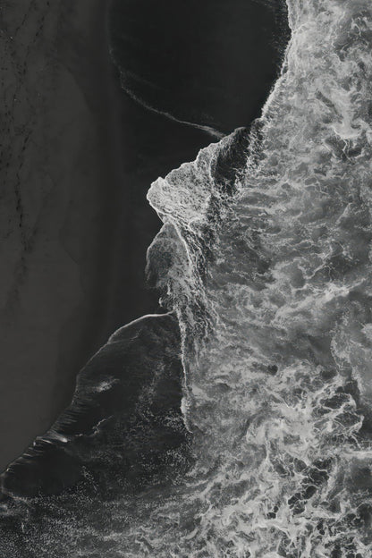 Black and white aerial photograph of ocean waves crashing against a dark rocky shoreline, with high-contrast textures of foam and sand.