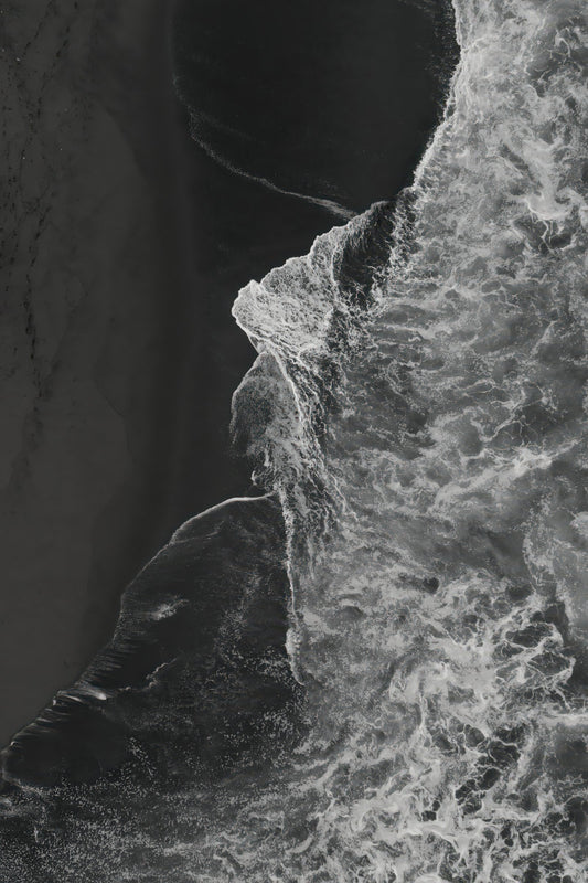 Black and white aerial photograph of ocean waves crashing against a dark rocky shoreline, with high-contrast textures of foam and sand.