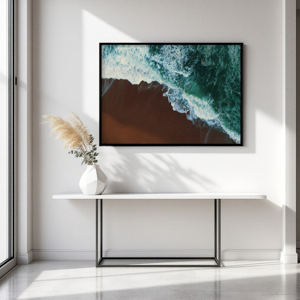 A framed wall art print showing an aerial view of turquoise ocean waves meeting a dark brown shoreline, displayed above a white console table with a vase in a bright modern room.