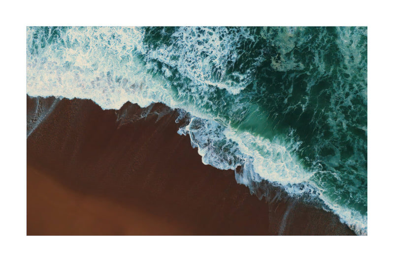 An aerial photograph of ocean waves with white foam crashing onto a dark brown sandy shore, presented as a square art print with a white border.