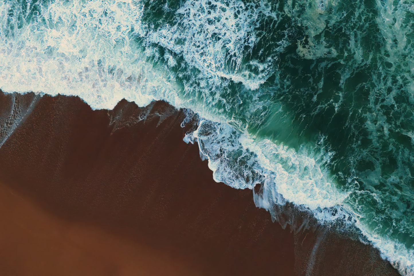 Aerial view of ocean waves with white foam crashing onto a brown sandy beach, filling the frame.