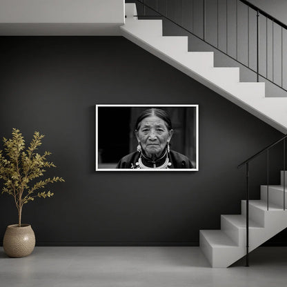 A black and white framed portrait print of an elderly woman with long hair and earrings, displayed on a dark gray wall in a modern interior setting with a staircase and a potted plant.