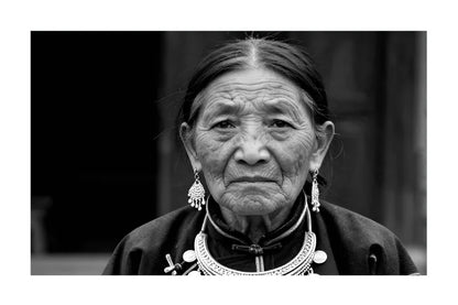 Black and white portrait photograph of an elderly woman wearing traditional jewelry and a high-neck garment, centered on a white background.