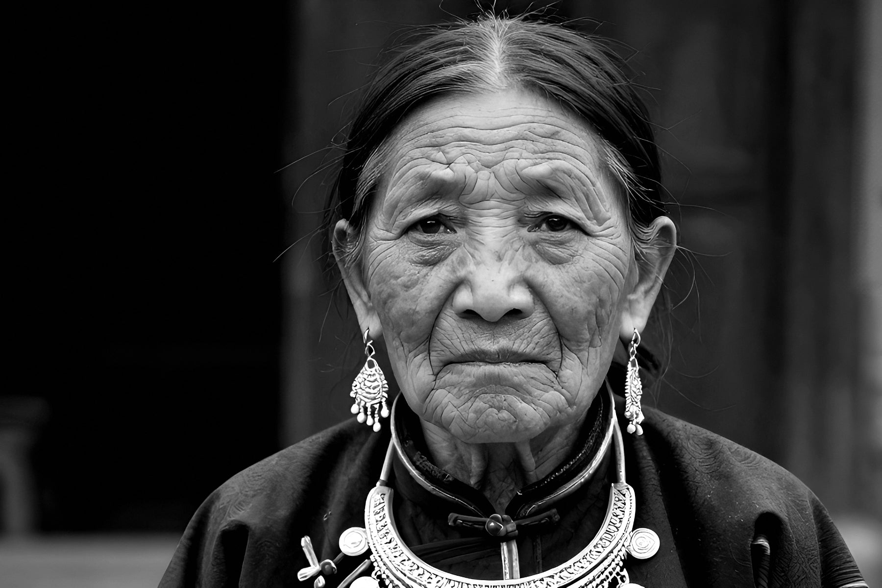 Black and white portrait of an elderly woman wearing traditional silver jewelry, including a large necklace, earrings, and a cross pendant, with a dark high-neck garment.