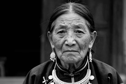 Black and white portrait of an elderly woman wearing traditional silver jewelry, including a large necklace, earrings, and a cross pendant, with a dark high-neck garment.