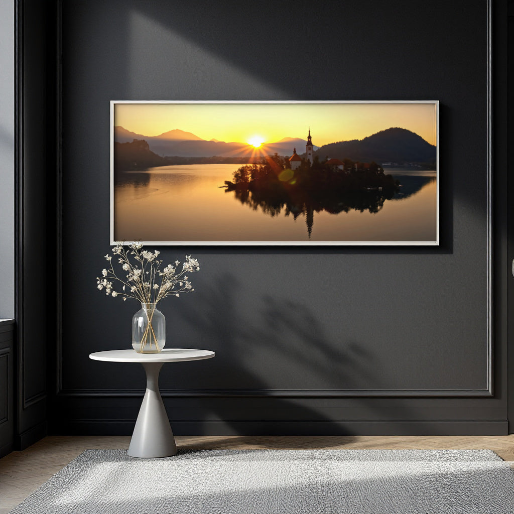 A framed landscape art print of a sunset over a lake with an island church and mountains, displayed on a dark charcoal wall above a small white side table with a vase of white flowers.