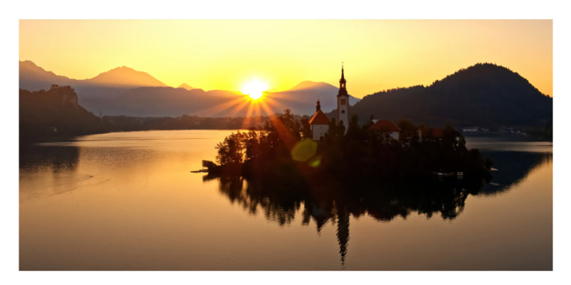A scenic landscape photograph of a small island church on a calm lake at sunset, with mountains in the background and the sun low on the horizon.