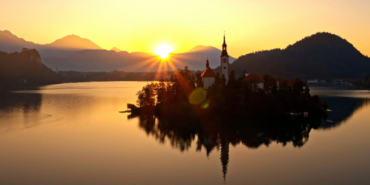 A scenic sunset landscape of an island with a church spire on a calm lake, surrounded by mountains and reflected in the water.
