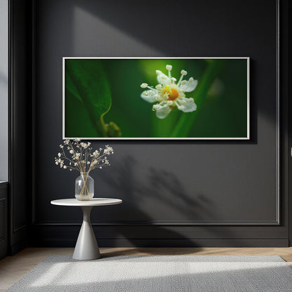 A framed wall art print showing a close-up white flower with a yellow center on a dark green background, displayed above a small round side table with a vase of dried stems against a dark charcoal wall.
