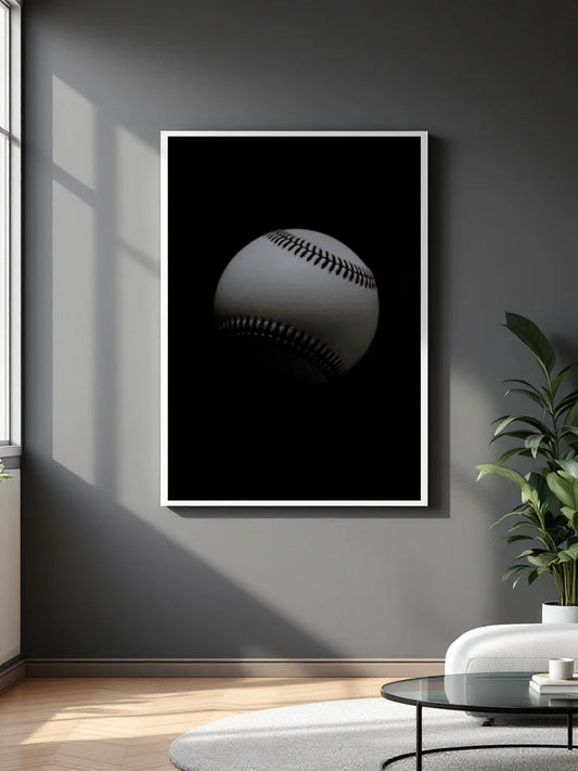 A vertical framed wall art print featuring a close-up baseball on a black background, displayed on a dark gray wall in a modern living room setting with a plant and white sofa.
