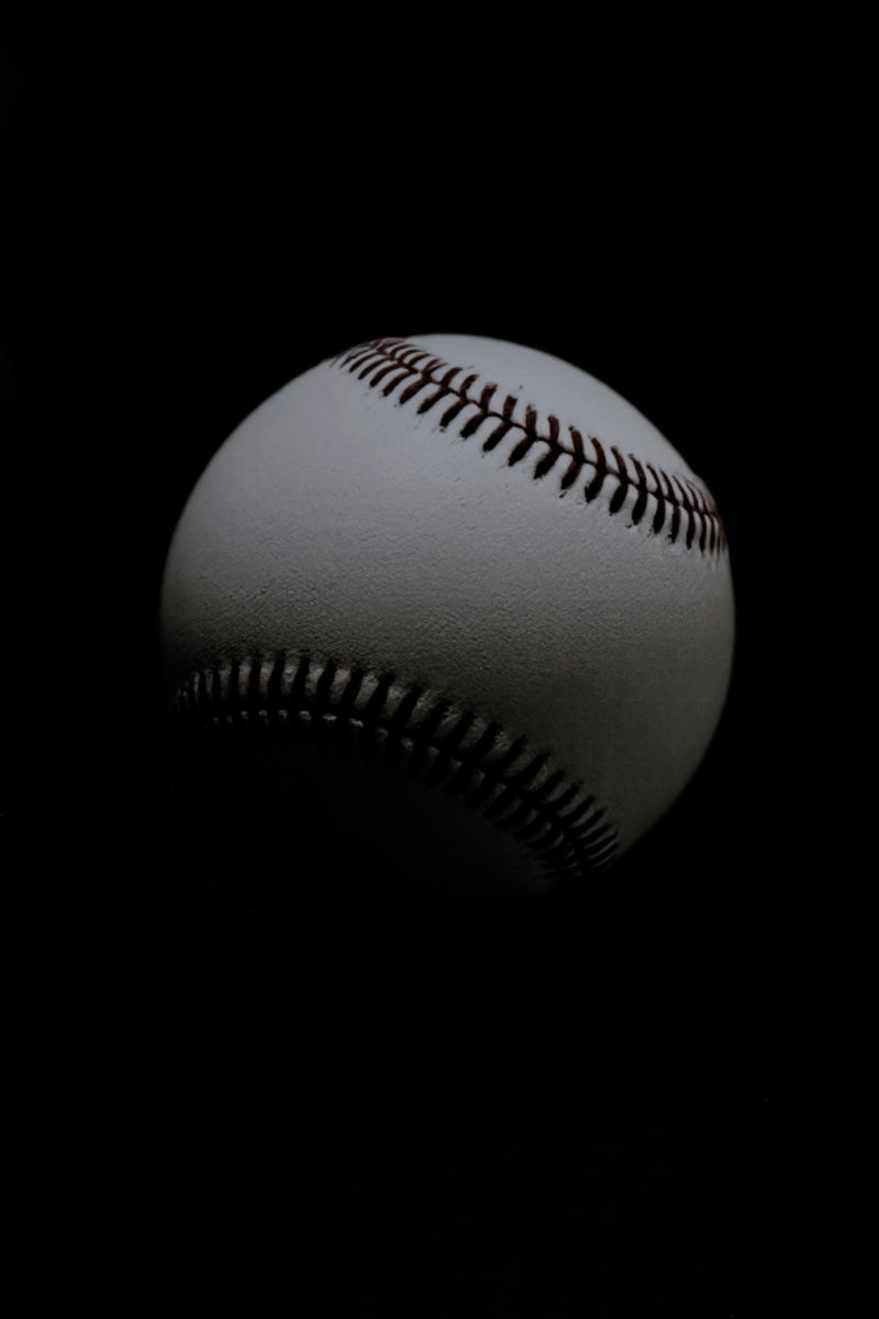 A close-up studio shot of a white baseball with dark red stitching against a solid black background.