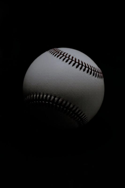 A close-up studio shot of a white baseball with dark red stitching against a solid black background.