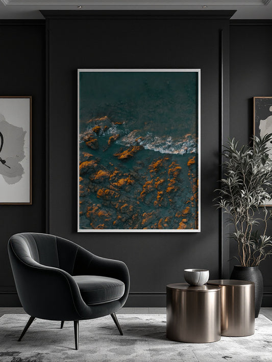 A framed vertical art print depicting an aerial view of dark teal water and golden-orange rocks, displayed on a dark charcoal wall in a modern living room setting with a black armchair and metallic side tables.