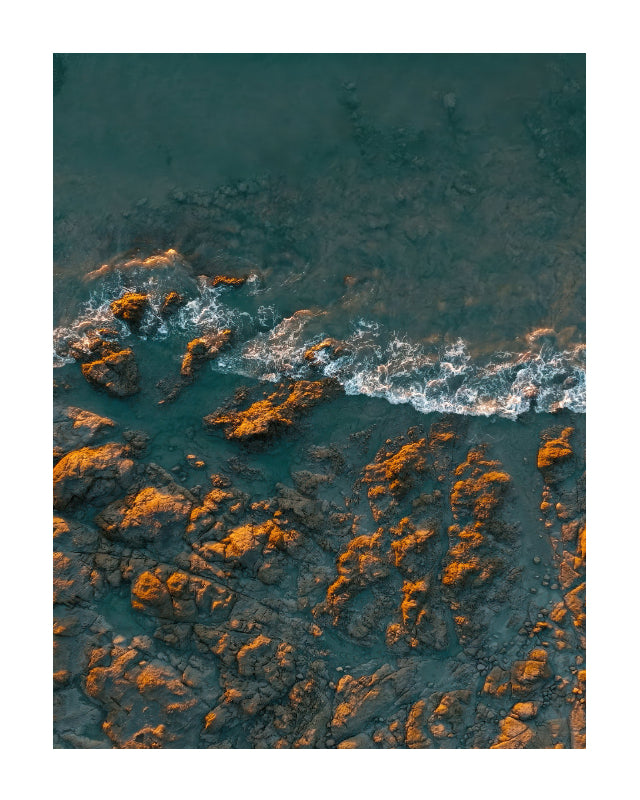 Aerial photograph of dark rocky shoreline with turquoise water and white surf, presented as a square art print with a white border.