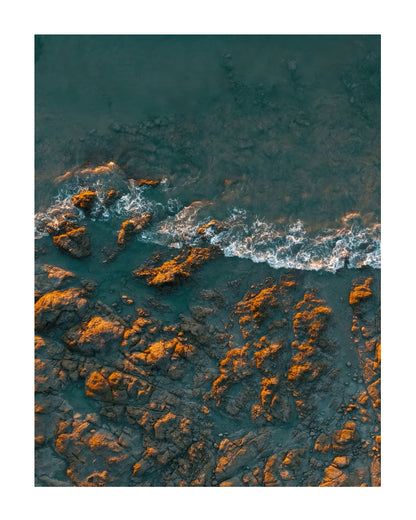Aerial photograph of dark rocky shoreline with turquoise water and white surf, presented as a square art print with a white border.