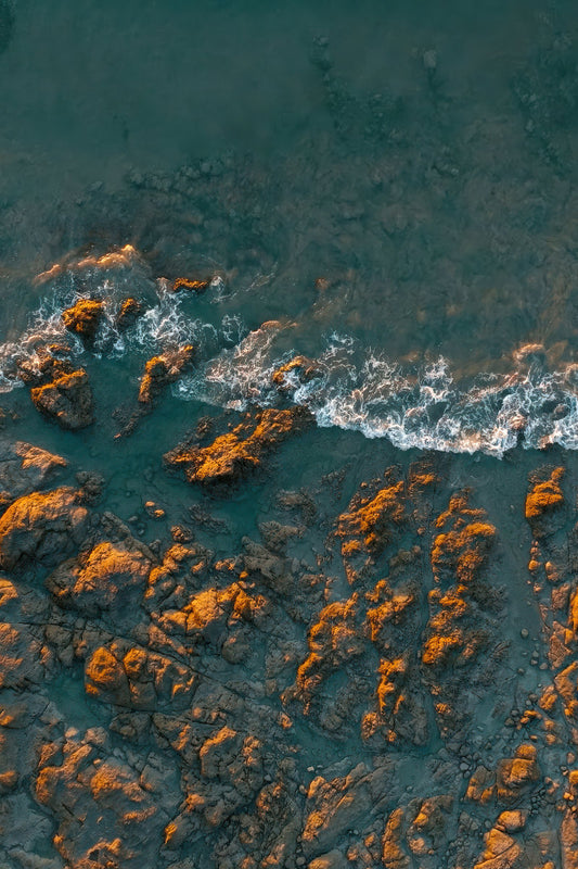 Aerial view of turquoise ocean water washing over rocky shoreline with warm orange-brown rocks.