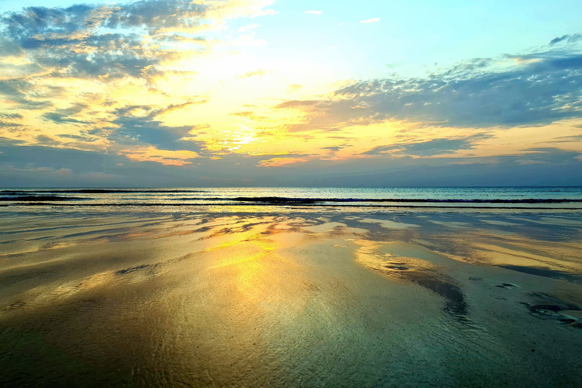 A scenic sunset over a calm sea with golden light reflecting on wet sand and water, under a cloudy sky.