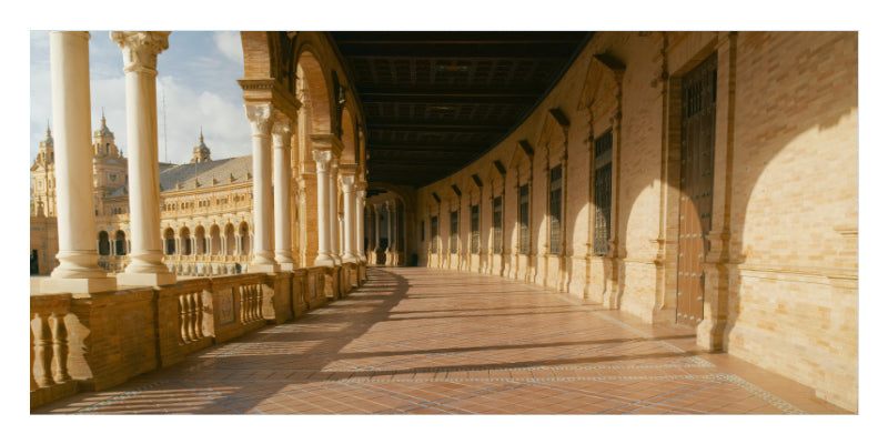 A long covered arcade walkway with repeating columns, warm beige stone walls, and a tiled floor, photographed in bright sunlight with strong shadows.
