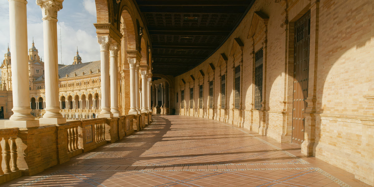 A long covered arcade walkway with repeating arches and columns, paved with reddish-brown tiles, set against warm beige stone walls under a dark ceiling. A large historic building is visible in the background through the left-side arches.