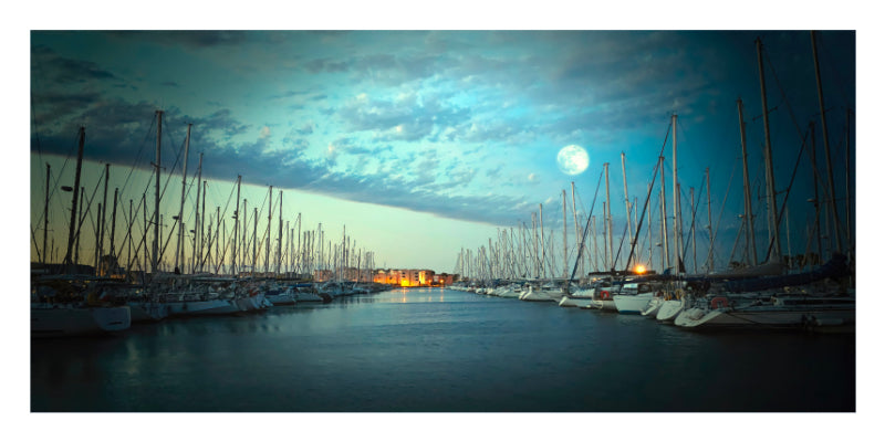 A scenic photograph of a marina at dusk or dawn under a full moon, with rows of sailboat masts lining a calm water channel and warm lights visible in the distance.