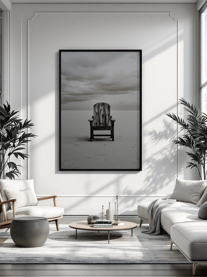 A framed black and white art print of a single wooden chair on a beach, displayed on a white wall in a modern, sunlit living room with a sofa, armchair, coffee table, and potted plants.