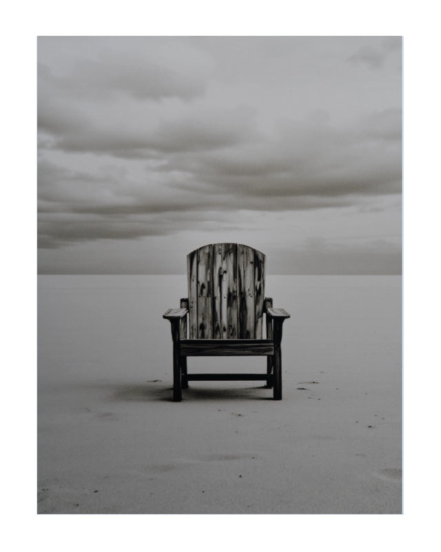 A weathered wooden Adirondack chair sits alone on a flat, pale beach under a cloudy gray sky.