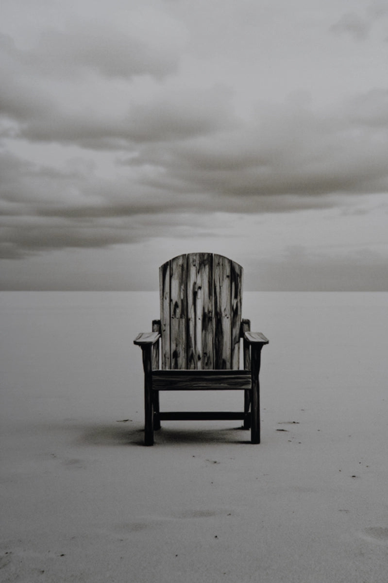 A single weathered wooden Adirondack-style chair sits centered on a flat, pale beach or sand dune under a cloudy gray sky.
