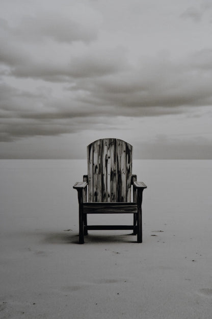 A single weathered wooden Adirondack-style chair sits centered on a flat, pale beach or sand dune under a cloudy gray sky.