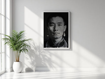 A framed black and white portrait print of a man with traditional earrings, displayed on a light gray wall in a bright room with a potted plant and window shadows.