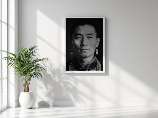 A framed black and white portrait print of a man with traditional earrings, displayed on a light gray wall in a bright room with a potted plant and window shadows.