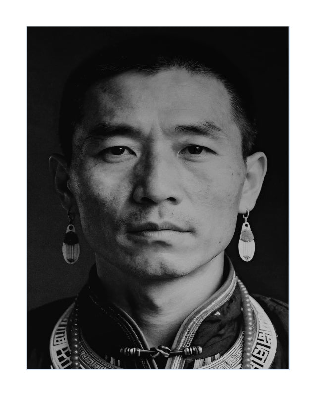 Black and white portrait photograph of a man wearing traditional ethnic jewelry, including a high collar with decorative trim and dangling earrings, set against a dark background with a white border.