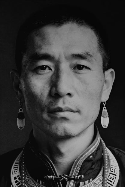 Black and white portrait of a man wearing traditional patterned clothing and long dangling earrings against a dark background.