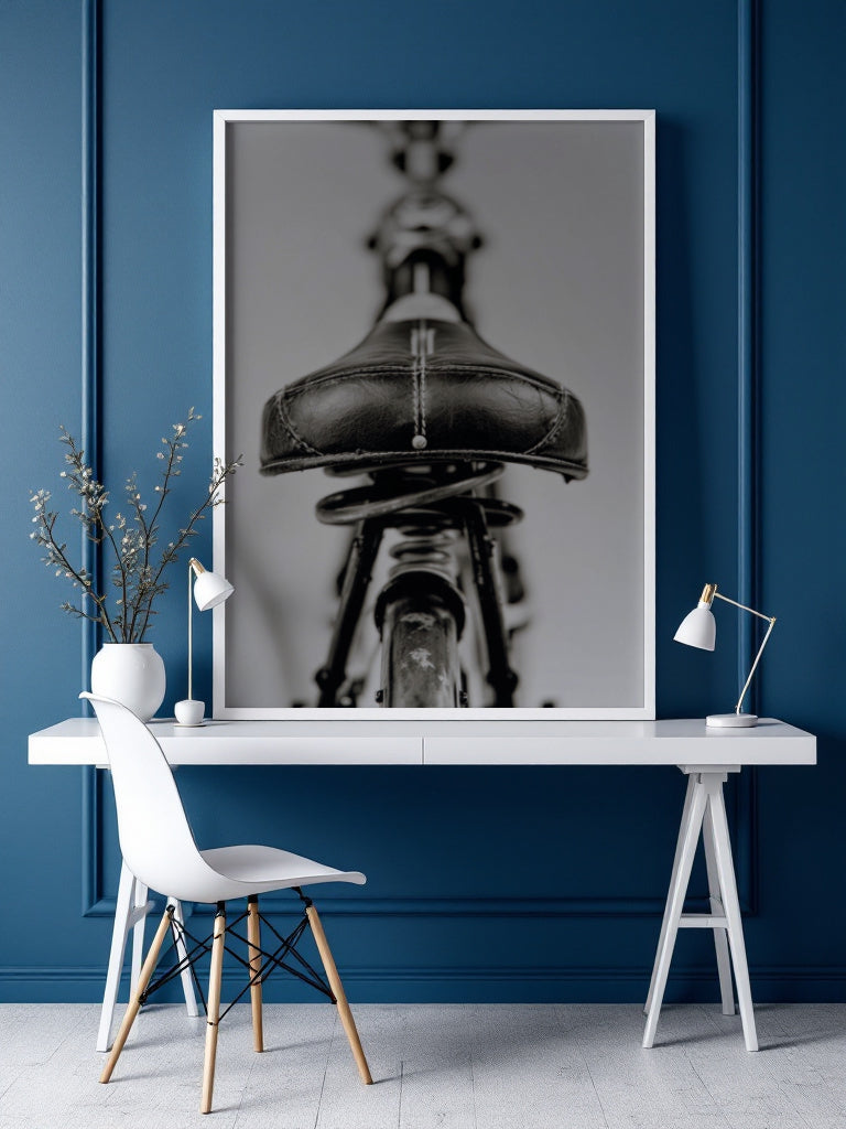 A large framed black and white art print of a bicycle seat hangs on a dark blue paneled wall above a white desk with a chair and lamps.
