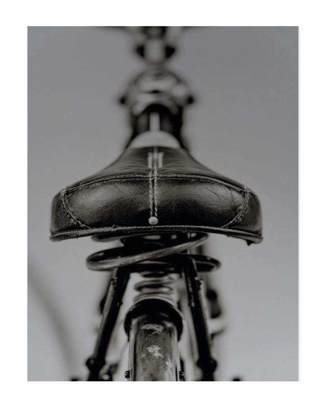 A close-up, low-angle photograph of a black leather bicycle saddle with a spring, shown in monochrome against a neutral gray background.