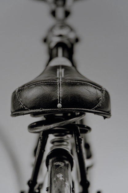 A close-up, low-angle view of a black leather bicycle saddle with white contrast stitching, mounted on a dark metal bicycle frame against a neutral gray background.