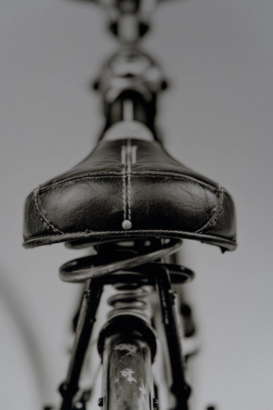A close-up, low-angle view of a black leather bicycle saddle with white contrast stitching, mounted on a dark metal bicycle frame against a neutral gray background.