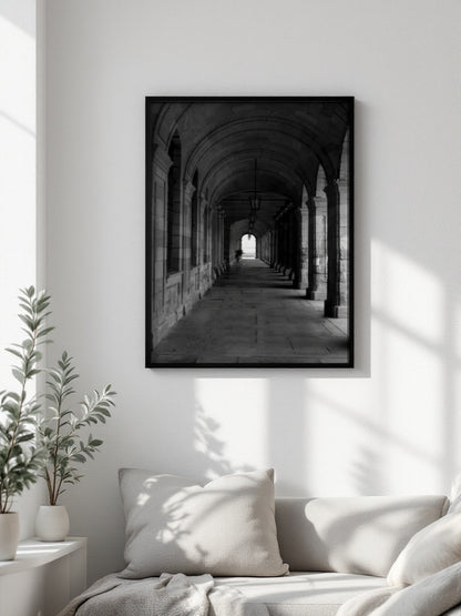 A black-framed black and white art print of a stone archway corridor, displayed on a white wall above a light-colored sofa with pillows in a bright, modern living room.