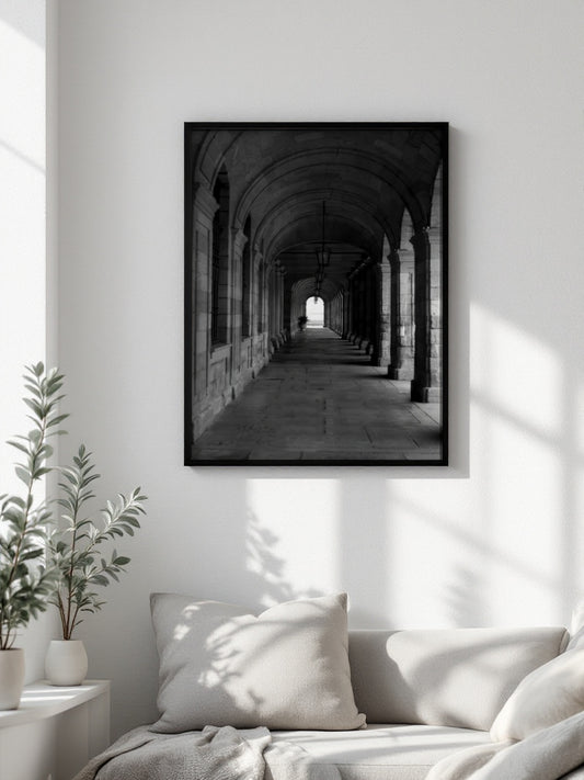 A black-framed black and white art print of a stone archway corridor, displayed on a white wall above a light-colored sofa with pillows in a bright, modern living room.