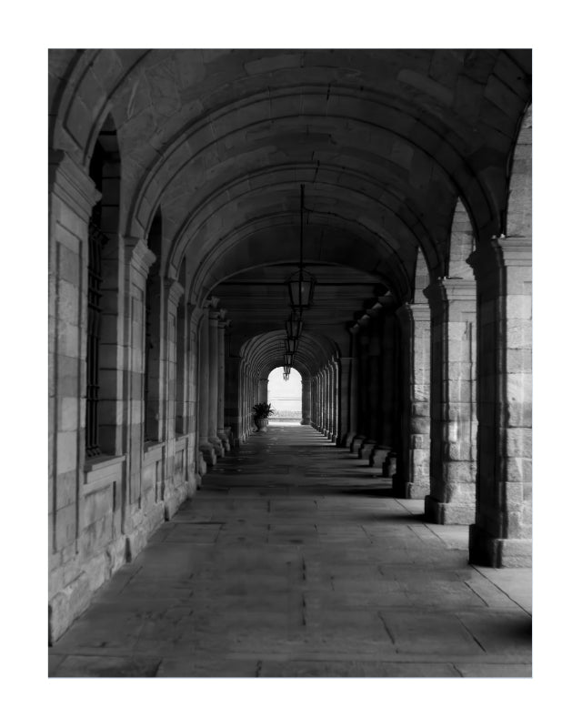 Black and white architectural photograph of a stone arcade corridor with repeating arches and columns leading into the distance.