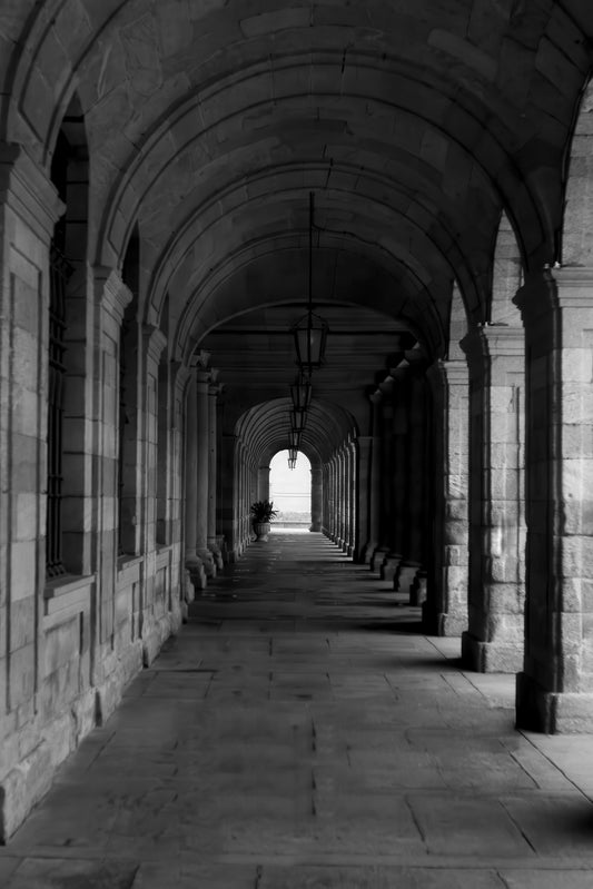 Black and white photograph of a stone colonnade with repeating arches and pillars leading the eye toward a bright opening at the end of the corridor.