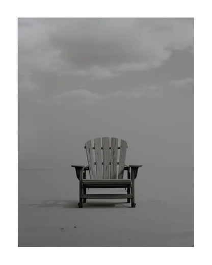 A single light-colored wooden Adirondack-style chair sits centered on a flat, misty gray surface under a cloudy overcast sky.