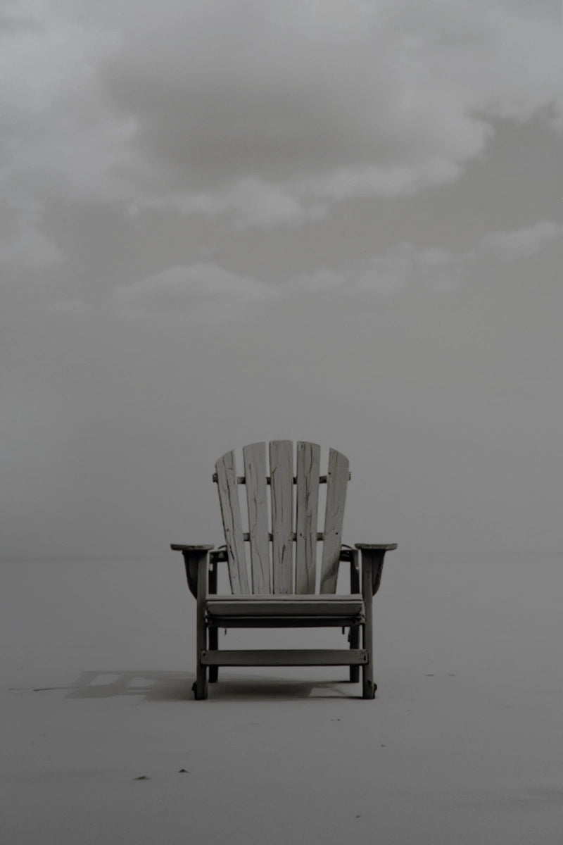 A single Adirondack-style chair centered on a flat, misty gray surface under a cloudy sky.