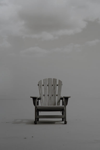 A single Adirondack-style chair centered on a flat, misty gray surface under a cloudy sky.