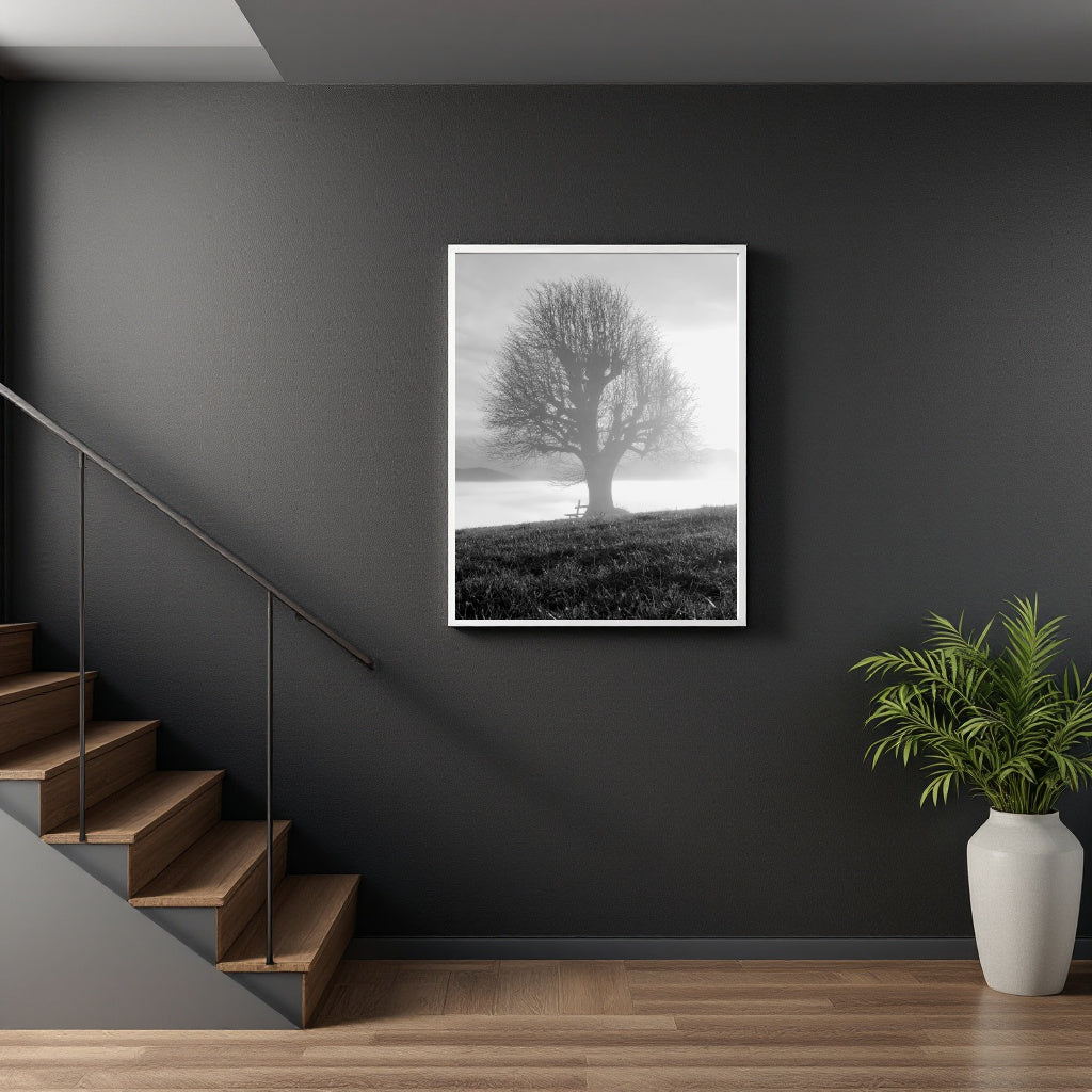 A framed black and white art print of a solitary tree in misty foggy terrain, displayed on a dark gray wall in a modern interior setting with a wooden staircase on the left and a potted green plant on the right.
