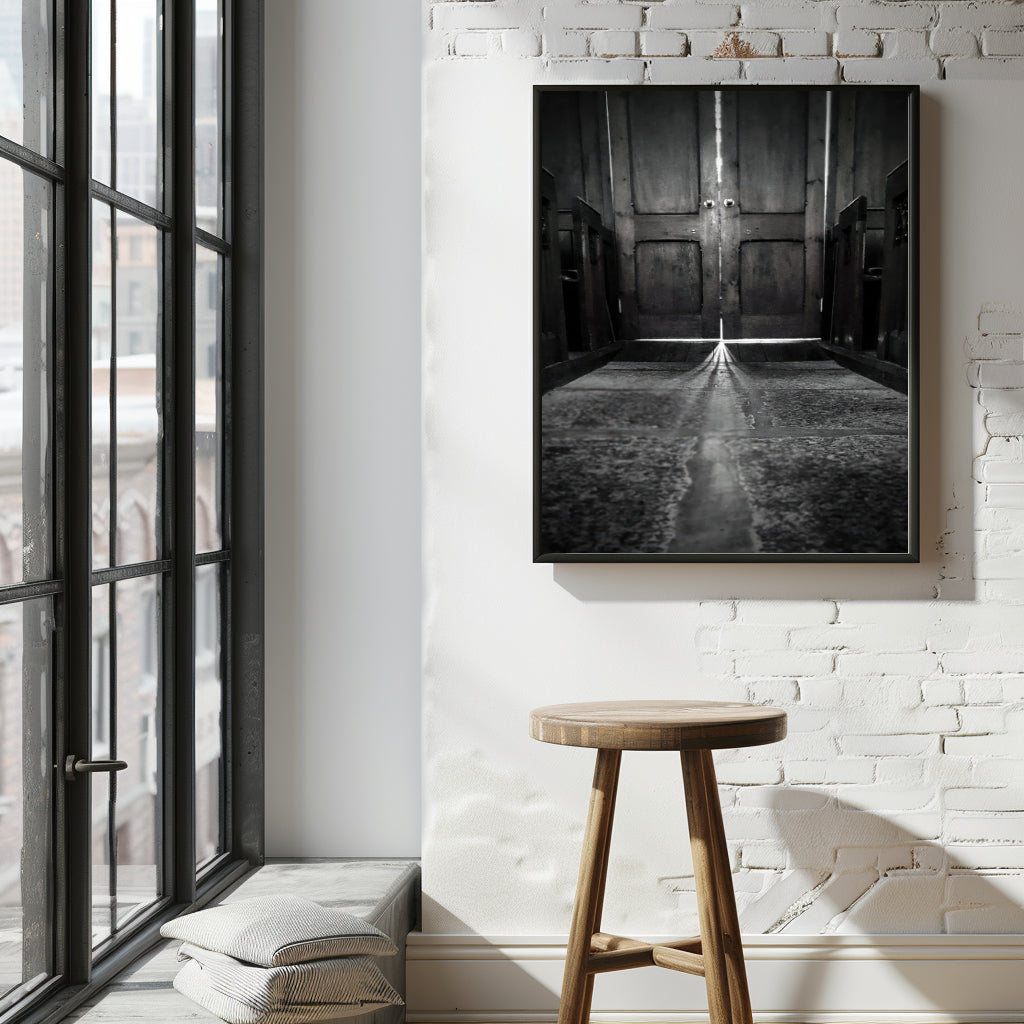 A black and white framed art print depicting a long hallway with a central staircase, displayed on a white brick wall above a wooden stool in a modern loft room setting.