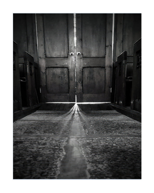 Black and white fine art photograph of a church interior looking up at a pair of double wooden doors with light shining in from the doorway.