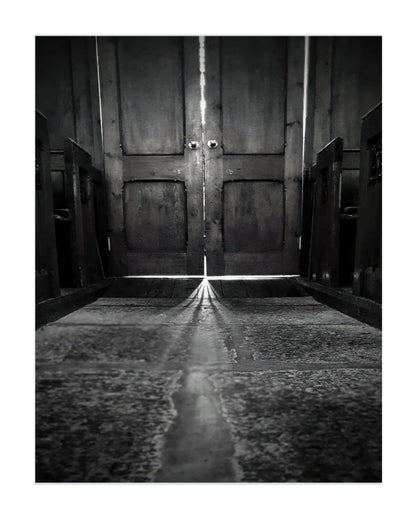 Black and white fine art photograph of a church interior looking up at a pair of double wooden doors with light shining in from the doorway.