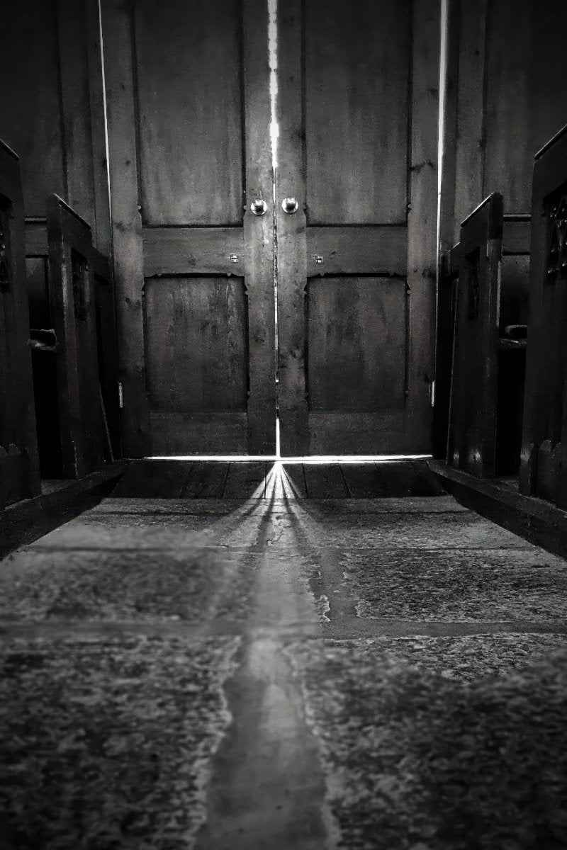 Black and white photograph of a dark wooden door with a bright beam of light shining through the center, casting a long reflection on a stone floor.