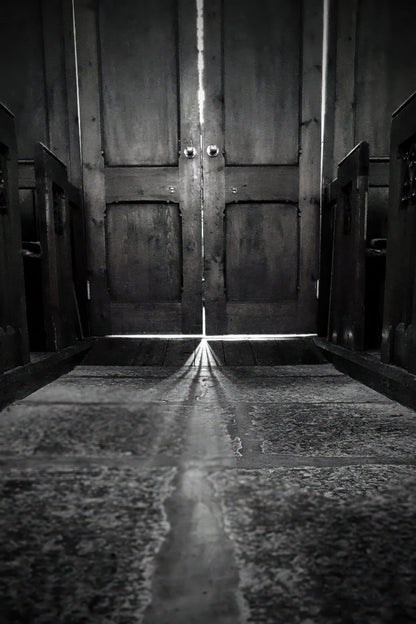 Black and white photograph of a dark wooden door with a bright beam of light shining through the center, casting a long reflection on a stone floor.