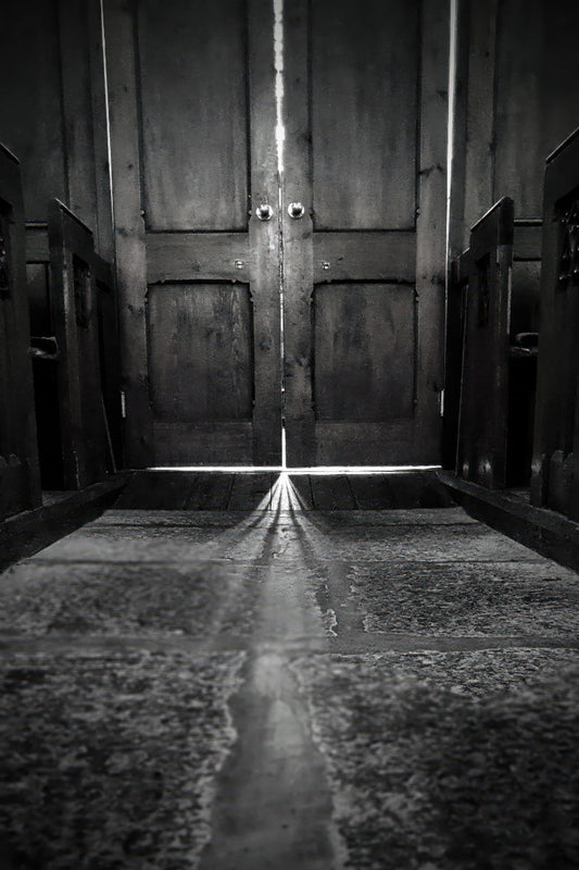 Black and white photograph of a dark wooden door with a bright beam of light shining through the center, casting a long reflection on a stone floor.